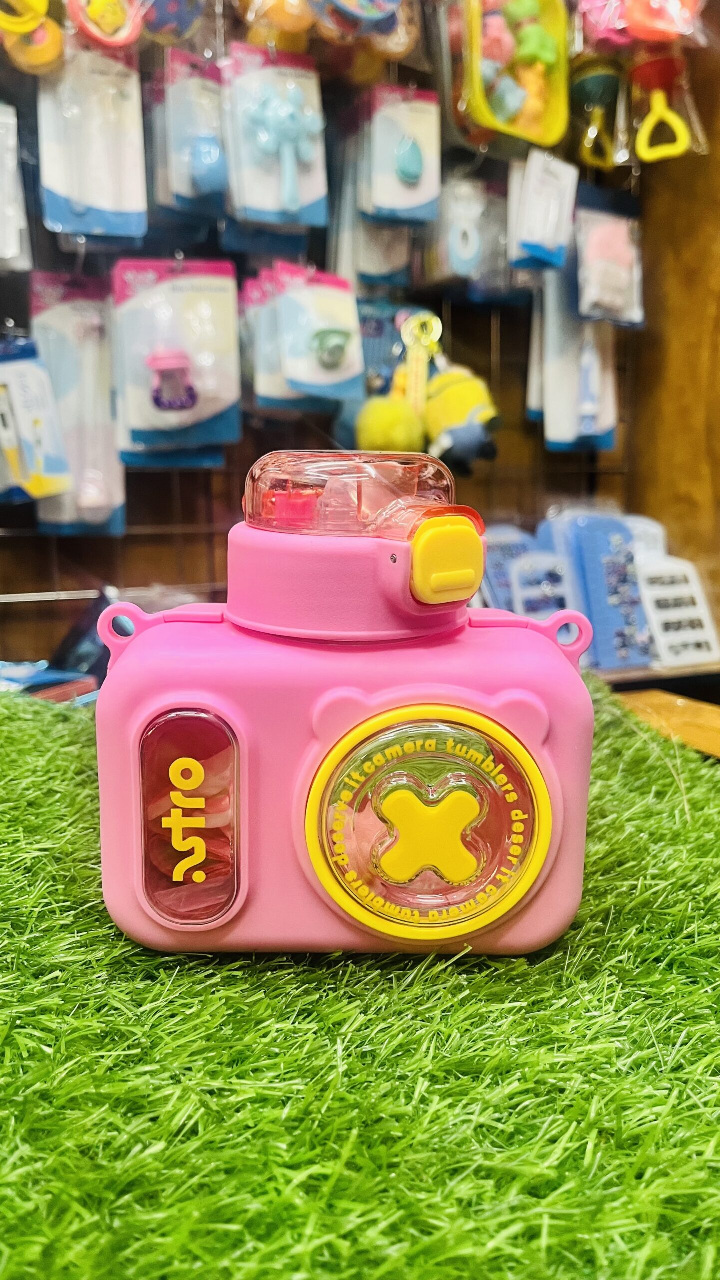 cute camera shape kids water bottle with silicone straw & shoulder strap (pink, 400 ml) Front view of closed cute camera shape kids water bottle with silicone straw in pink standing on green grass