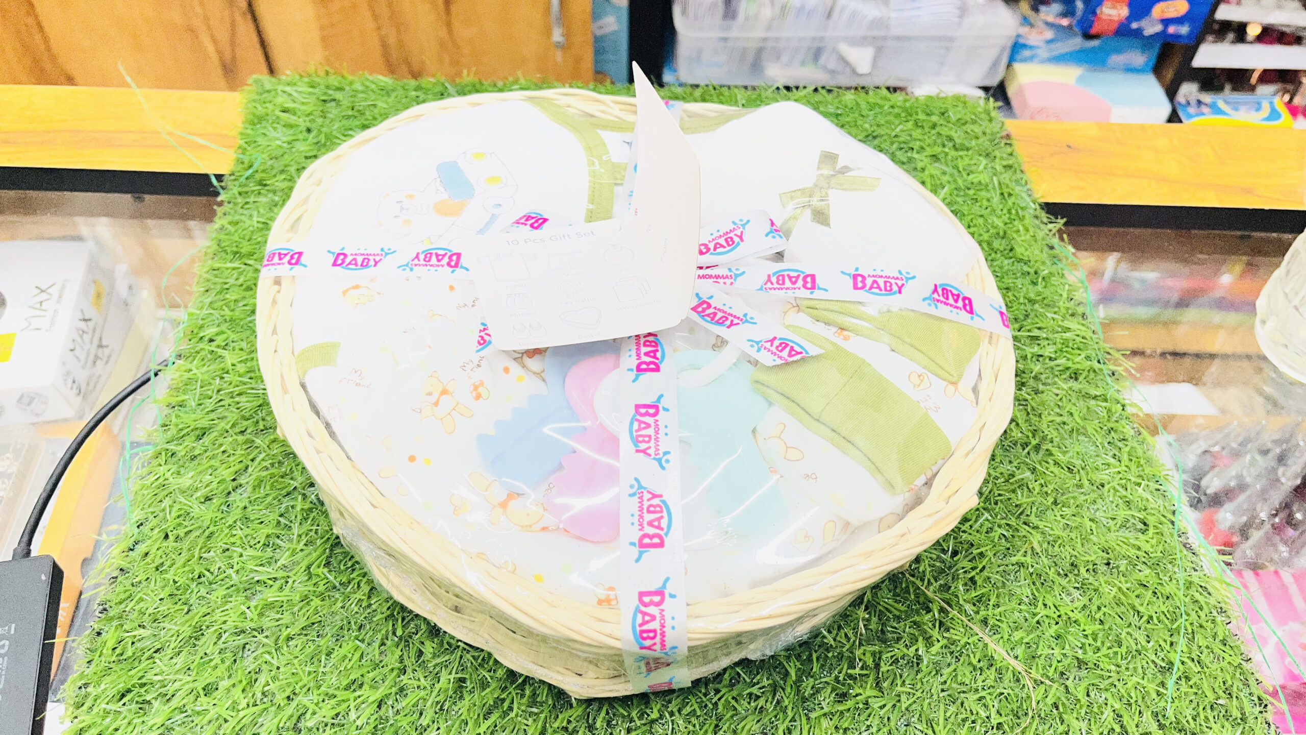 mommas baby 10 pcs newborn baby gift heart basket set – 100% cotton with rattle (sage green) Top down view of Mommas Baby 10 pcs newborn baby gift heart basket set wrapped in clear film with ribbon on grass